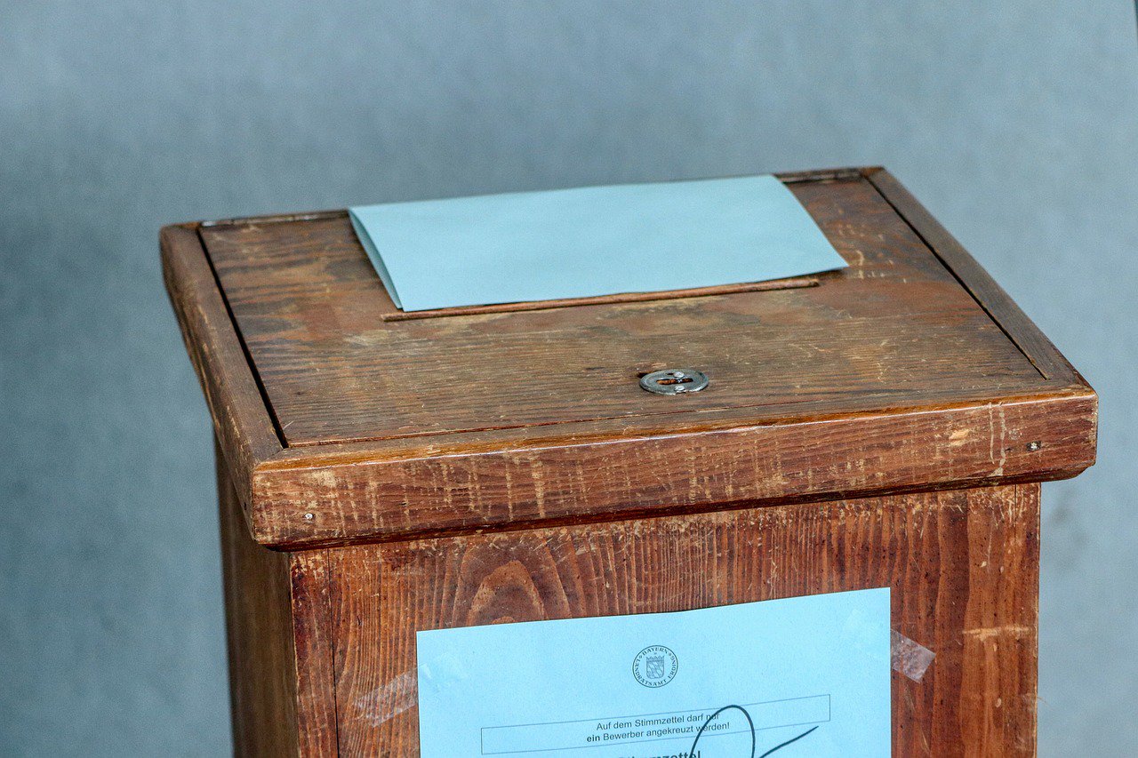 A wooden ballot box with an envelope partly inserted in the slot.