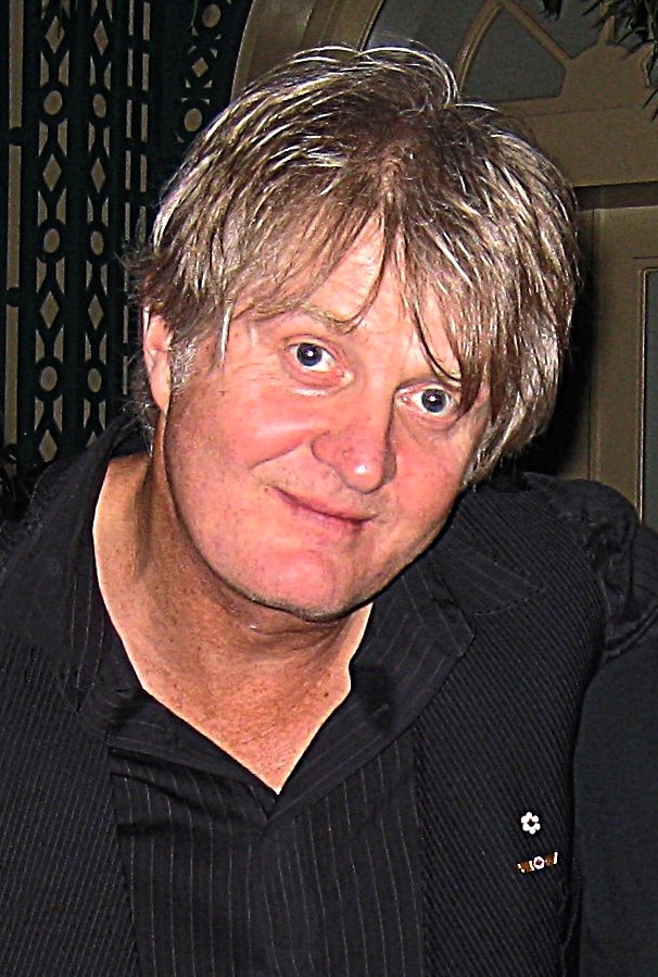 Tom Cochrane smiling at the camera, wearing a dark pinstriped jacket over a black v-neck shirt.