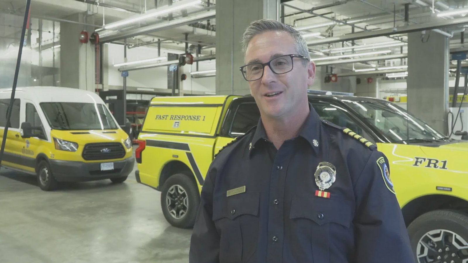Victoria Fire Chief Dan Atkinson speaking in a fire hall garage, with emergency response vehicles in the background.